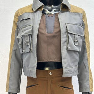 May include: A gray jacket with tan accents, featuring multiple front pockets and a brown undershirt. The jacket has a collar and is paired with brown pants. The outfit is displayed on a mannequin.