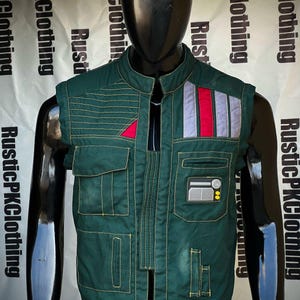 Peut inclure: Un gilet vert foncé avec plusieurs poches et des coutures jaunes. Le gilet présente un triangle rouge et un patch avec des rayures grises, rouges et blanches. Un mannequin noir présente le gilet, mettant en valeur son design et ses détails.