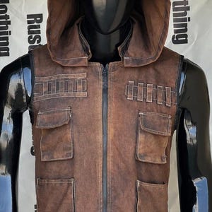 Peut inclure: Gilet à capuche marron avec plusieurs poches et une fermeture éclair sur le devant. Le gilet a un aspect vieilli avec des détails de couture visibles. La capuche est attachée et le gilet est présenté sur un mannequin noir.