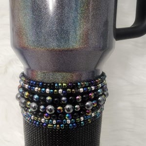 May include: A black and silver tumbler with a black handle. The tumbler is decorated with a band of black, silver, and iridescent beads.