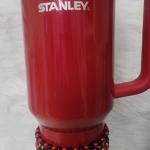 May include: A red Stanley travel mug with a black and red plaid fabric band and a set of beaded bracelets around the mug.