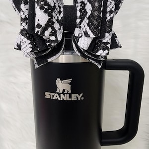 May include: A black Stanley tumbler with a handle, topped with a large bow. The bow features a black and white snakeskin pattern. The tumbler has the Stanley logo in white.