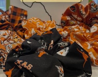 May include: A collection of scrunchies in various patterns, including orange and white, and black with orange accents. The scrunchies feature the text "THE UNIVERSITY OF TEXAS" and the word "TEXAS". The background includes the words "SIZE SCRUNCHIES".
