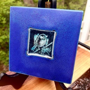 May include: A square, cobalt blue ceramic tile with a detailed bird illustration in a smaller, inset square. The bird is perched on a branch with turquoise accents. The tile is displayed on a black easel, showcasing the artwork.