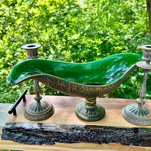 May include: A centerpiece bowl with a glossy emerald green interior and a textured, weathered bronze-toned exterior, flanked by two matching metal candlesticks featuring a twisted stem design and a wide, circular base with decorative detailing.
