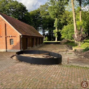 May include: An outdoor scene featuring a brick building with a red tile roof and multiple brown doors. A black circular structure is in the foreground, surrounded by a brick paved area. Trees and greenery fill the background.