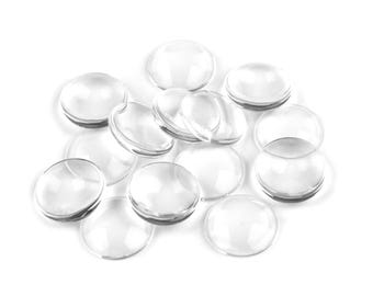 May include: A group of clear glass cabochons, also known as dome-shaped glass beads, are scattered on a white surface.