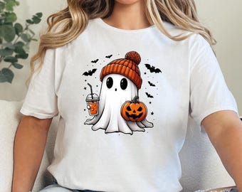 May include: White t-shirt with a cartoon ghost wearing an orange and white hat, holding a pumpkin and a cup of orange juice. The ghost is surrounded by bats and stars.