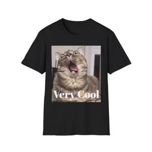 May include: A black short-sleeved t-shirt featuring a square graphic print of a brown tabby cat with its mouth wide open and tongue visible. Below the cat, the text "Very Cool" is printed in a white serif font centred on the graphic.