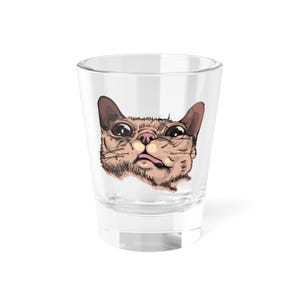 May include: Clear shot glass with a cartoon cat face design. The cat has big eyes, a pink nose, and its tongue is out. The illustration uses brown, tan, and pink colours. A fun and quirky item.