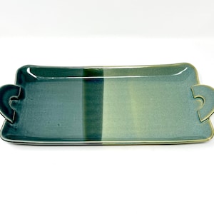 May include: A rectangular serving tray with two handles on the sides. The tray is made of ceramic and has a green and blue glaze. The glaze is uneven, creating a mottled effect.