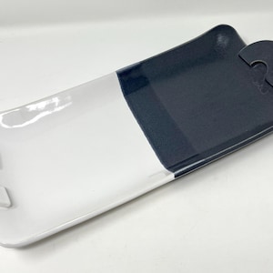 May include: A rectangular ceramic serving tray with a two-tone design. One half is white, the other is dark blue. Each end has a curved handle. The tray is suitable for serving appetizers or small dishes.