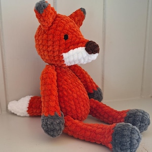May include: A handmade plush fox toy in vibrant orange, white, brown, and grey. The fox features a fluffy texture, with a white muzzle, a brown nose, and grey accents on the ears and feet. The fox is sitting with its legs extended.