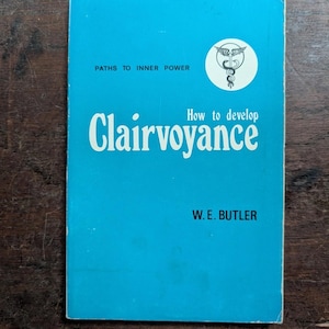 May include: A vintage book with a turquoise cover titled "How to develop Clairvoyance" by W.E. Butler. The cover also features the text "Paths to Inner Power" and a circular emblem. The book is set against a wooden surface.
