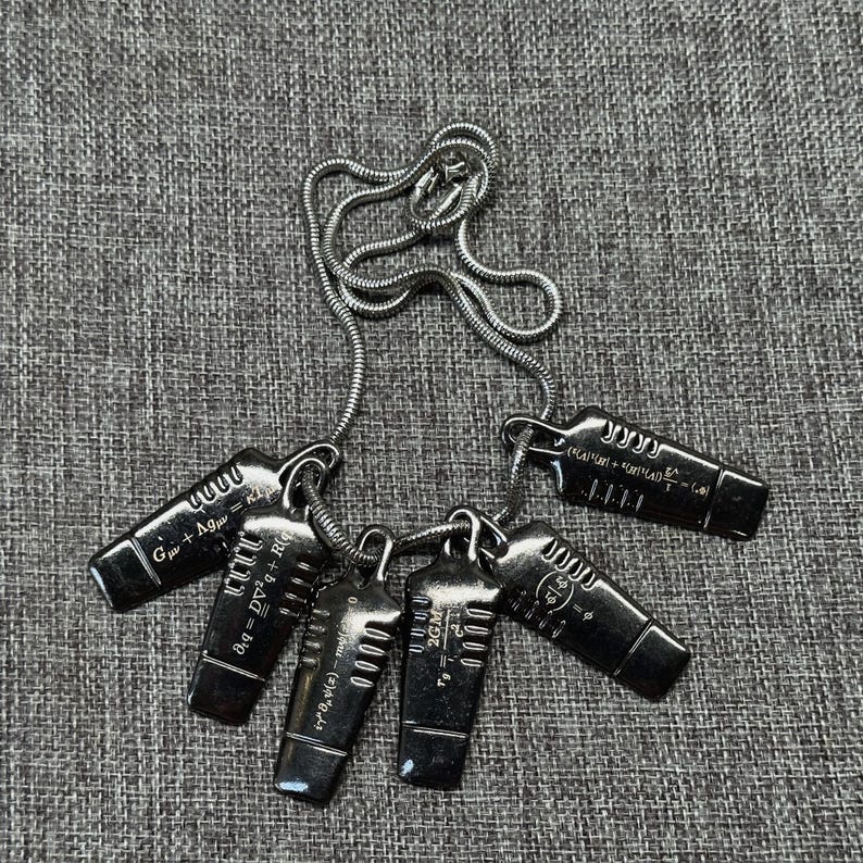 May include: A silver-toned necklace featuring five rectangular pendants. Each pendant has engraved scientific formulas and the number "0000". The chain is a delicate, coiled design.