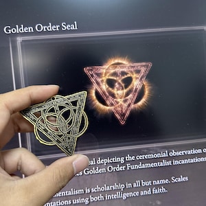 May include: A gold-colored metal pendant with a triangular design featuring a complex pattern of overlapping circles and lines. The pendant is similar to a symbol seen in the video game Elden Ring, known as the Golden Order Seal.