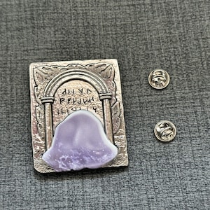 May include: A silver-toned metal pin featuring a detailed archway with runes and a purple stone. The pin is attached to a backing with a safety pin.
