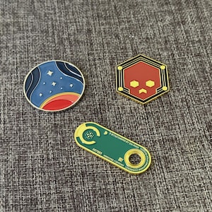 Puede incluir: Tres pines esmaltados con diseños únicos. Uno es una escena celeste azul y roja, otro es una calavera roja en un hexágono dorado, y el último es una etiqueta verde con texto y un agujero circular. Ideales para chaquetas, bolsos o colecciones.