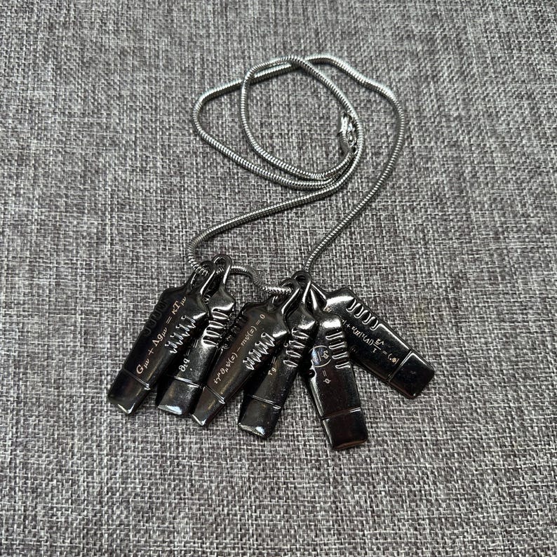 May include: A silver-toned necklace featuring six rectangular pendants. Each pendant has a unique design with etched lines and mathematical equations. The necklace chain is a thin, coiled design. The pendants are arranged on a gray fabric surface.