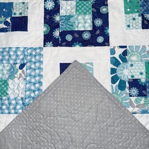 Puede incluir: Un edredón hecho a mano con un diseño de patchwork con cuadrados de patrones florales blancos, azules y turquesas. La parte inferior del edredón es gris con un patrón de lunares blancos y acolchado floral. El edredón está doblado, revelando el reverso gris.