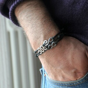 May include: A black leather bracelet with a silver charm featuring a stylized design of multiple loops.