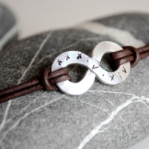 Peut inclure: Un bracelet en cuir marron avec un charme en argent en forme de symbole infini. Le charme est gravé de chiffres romains et d'autres marques.