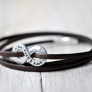 Op de afbeelding: Een bruin lederen armband met een zilveren oneindigheidsbedel met de tekst "to the moon & back".