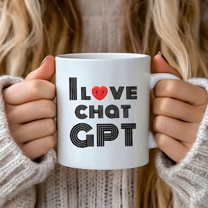 Puede incluir: Taza de cerámica blanca con el texto "I LOVE CHAT GPT" en negro, con un icono de corazón rojo. La taza es sostenida por una persona que lleva un suéter color crema.