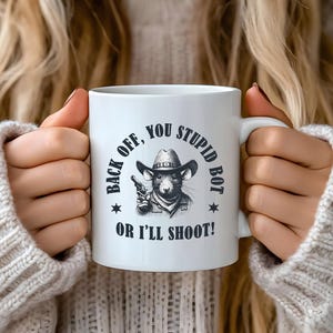 Op de afbeelding: Witte keramische mok met een humoristisch ontwerp. De mok heeft een cartoonrat met een cowboyhoed en een pistool, met de tekst "BACK OFF, YOU STUPID BOT OR I'LL SHOOT!" in het zwart gedrukt. De mok wordt door een persoon vastgehouden.