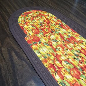 May include: Oval-shaped braided rug with a brown border. The rug's center features a repeating pattern of apples in shades of red, yellow, and green. The apples have green leaves and are stitched together with a zig-zag pattern.