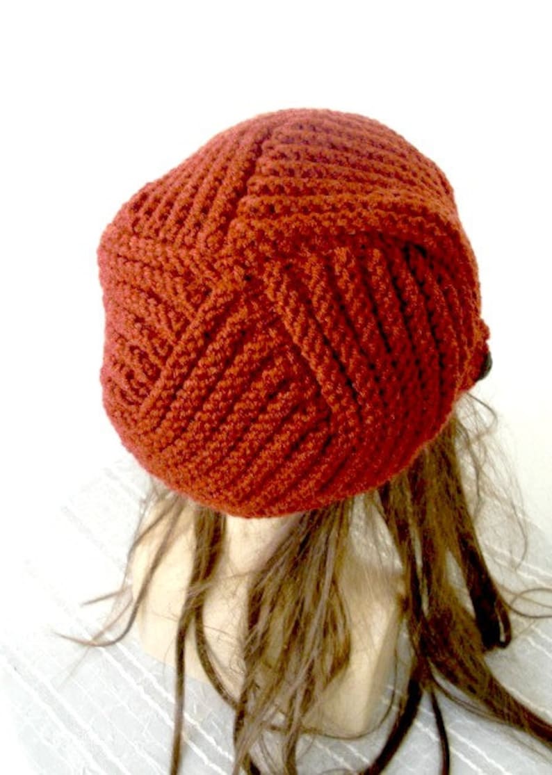 May include: A rust-colored crocheted beanie hat with a textured pattern. The hat is on a mannequin head with long brown hair.