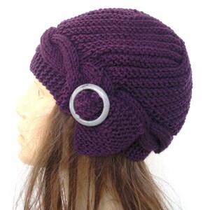 May include: A purple knitted hat with a decorative silver ring on the side. The hat is worn on a mannequin with long brown hair.
