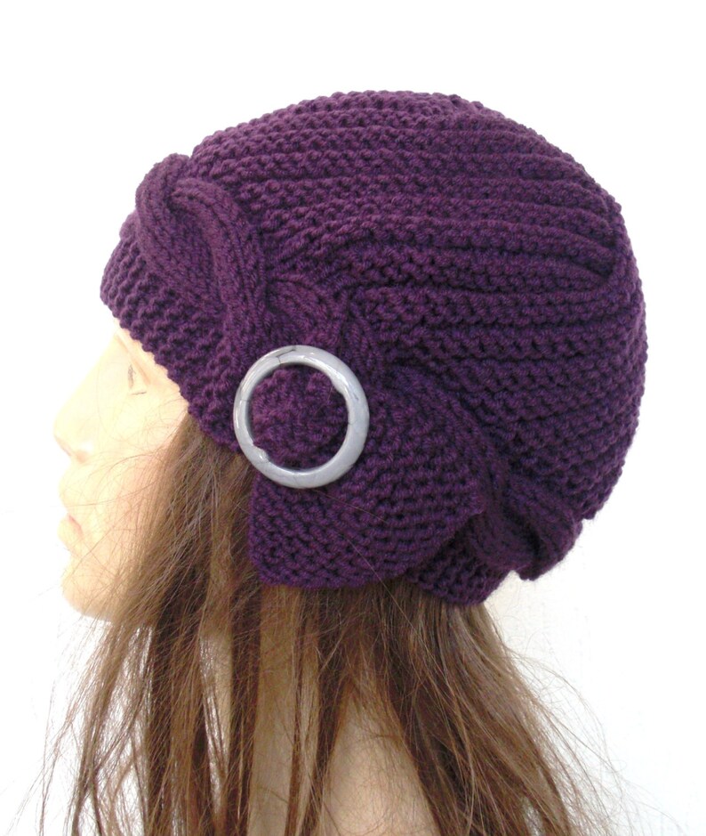 May include: A purple knitted hat with a decorative silver ring on the side. The hat is worn on a mannequin with long brown hair.