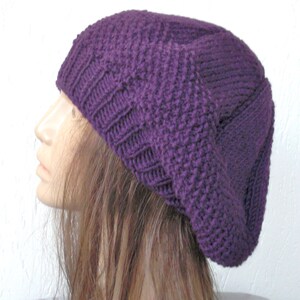 May include: A purple knitted beanie hat with a slouchy fit. The hat is made of a soft, textured yarn.