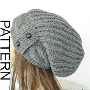 Puede incluir: Boina gris holgada con un patrón de punto de canalé y dos botones negros en el lateral.