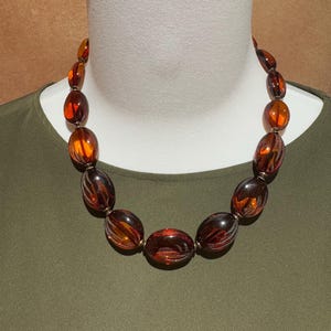 May include: A necklace featuring a series of polished, oval-shaped amber-coloured beads. The beads exhibit a translucent, deep orange and brown hue with visible internal striations, connected by small metallic spacers in a graduated arrangement.