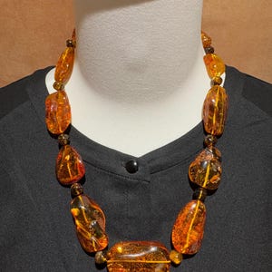 May include: A statement necklace featuring large, irregular amber beads. The beads are a warm, translucent orange colour. The necklace is displayed on a black garment.