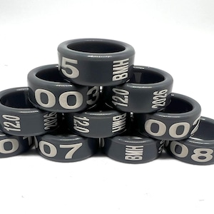 Peut inclure: Une pile d'anneaux en plastique gris foncé, chacun avec des chiffres et du texte blancs imprimés. Les anneaux affichent des chiffres tels que 00, 03, 05, 07 et 08, ainsi que "BMH", "12.0" et l'année "2026".