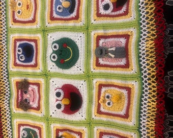 May include: A colorful, handmade crochet blanket featuring a grid of squares, each with a different cartoon character face. The blanket has a white, green, yellow, and red color scheme, with a decorative border.