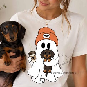 May include: A white t-shirt featuring a graphic of a white ghost wearing an orange beanie with a bat icon, holding a brown coffee cup and a black and tan dachshund puppy. The text DUONGVIVIDVERSE is printed in small black letters near the ghost.