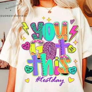 Peut inclure: T-shirt crème avec un graphisme coloré qui dit "YOU GOT THIS" dans différentes polices et couleurs. Le design comprend des éclairs, des cœurs et le hashtag "#testday". Le t-shirt est une pièce décontractée et motivante.