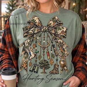 May include: A sage green t-shirt featuring a large camouflage print bow graphic. Hanging from the bow are illustrations of deer antlers, a mallard duck, binoculars, a shotgun shell, a tree, and a boot. The text Hunting Season is printed below.
