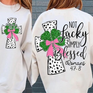 Puede incluir: Sudadera blanca con un diseño de cruz en blanco y negro que presenta un trébol de cuatro hojas verde con purpurina rosa y un lazo rosa. El texto "Not Lucky Simply Blessed Romans 4:7-8" está impreso en la sudadera.
