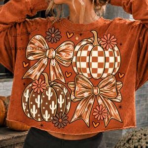 Può includere: Una felpa a maniche lunghe color arancio bruciato con una stampa grafica di due grandi fiocchi, due zucche e piccoli fiori. Una zucca presenta un motivo a scacchi bianchi, l'altra una stampa di cactus bianchi. Inclusi piccoli cuori e stelle.