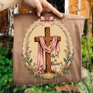 May include: A brown pillow with an oval design featuring a wooden cross draped with a pink cloth. The words "He is Risen" are below the cross. The design is surrounded by a floral wreath and a pink bow.