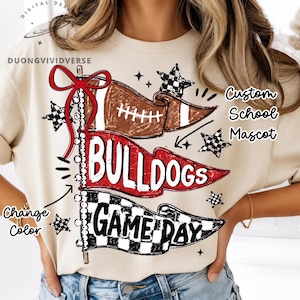 May include: A beige t-shirt featuring a graphic of three pennant flags on a pole with a red bow. The flags display a football, the text "BULLDOGS", and "GAME DAY" in a checkered pattern. Text includes "DUONGVIVIDVERSE", "Custom School Mascot", and "Change Color".