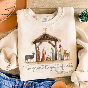 May include: A cream-colored sweatshirt with a watercolor illustration of the nativity scene. The illustration features a wooden manger, a star, and figures of Mary, Joseph, and baby Jesus. The text "The greatest gift of all" and "Matthew 1:21" is printed below the illustration.