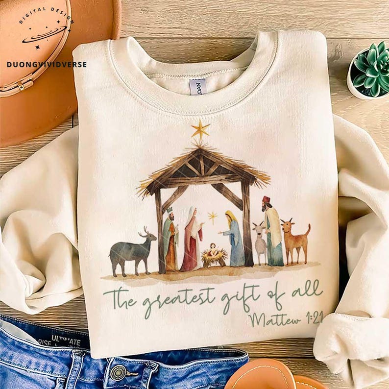 May include: A cream-colored sweatshirt with a watercolor illustration of the nativity scene. The illustration features a wooden manger, a star, and figures of Mary, Joseph, and baby Jesus. The text "The greatest gift of all" and "Matthew 1:21" is printed below the illustration.