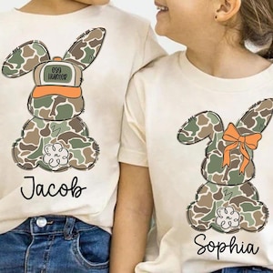 May include: Two white t-shirts with a camouflage bunny design. One bunny is wearing a hat that says "Egg Hunter". The shirts are personalized with the names "Jacob" and "Sophia".