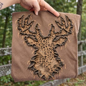 May include: A brown t-shirt featuring a large, distressed deer head silhouette appliqué. The deer shape is filled with a brown and black leopard print pattern, and the edges of the fabric are frayed to create a textured, raw-edge appearance.